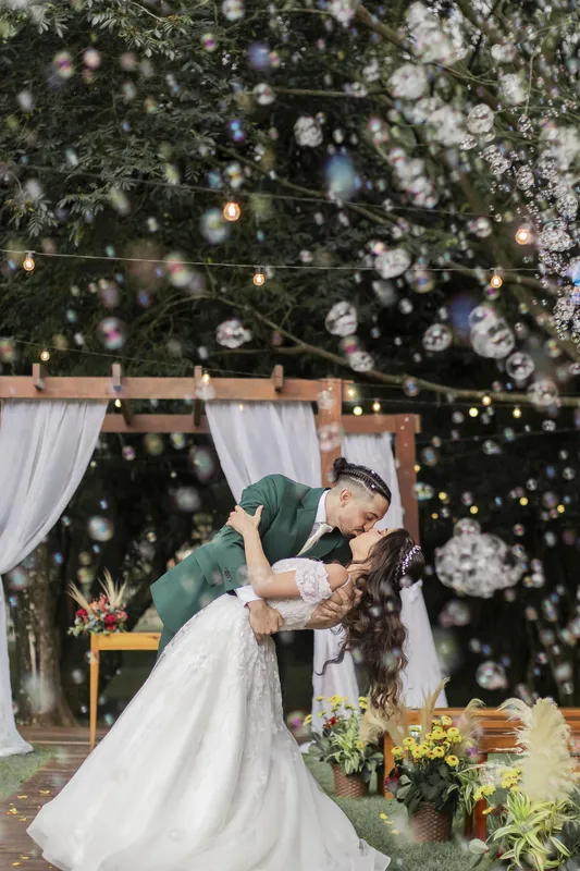Noivo inclinando a noiva para um beijo sob bolhas de sabão e luzes