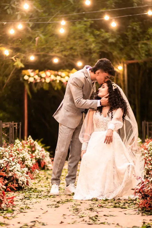 Noivo beijando a testa da noiva sentada no corredor de flores iluminado
