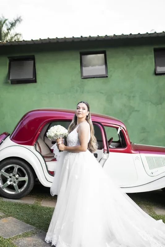 Noiva ao lado de um carro antigo vermelho em frente a uma casa verde