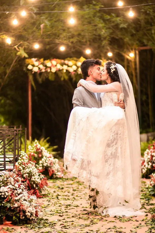 Noivo carregando a noiva e os dois se beijando sob um varal de lâmpadas