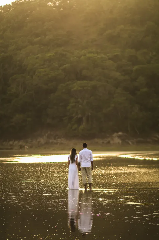 Casal de mãos dadas de costas dentro da água com o reflexo dourado