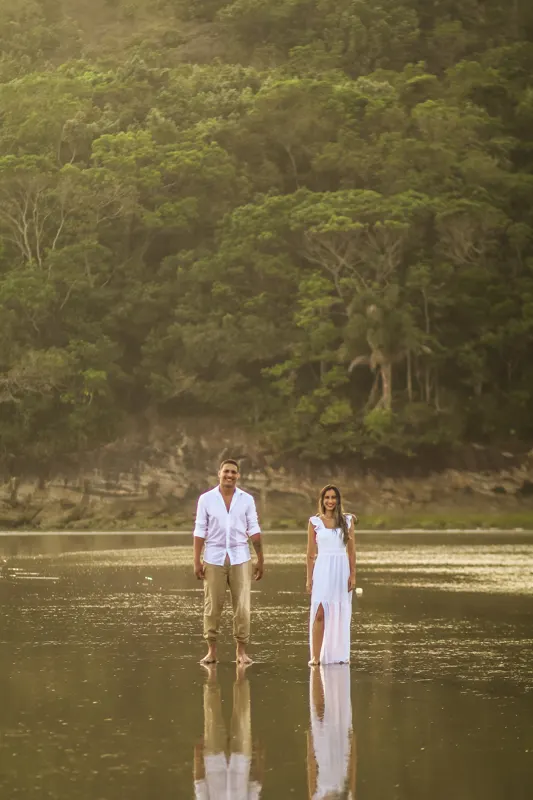 Casal sorrindo em pé na água com a mata ao fundo