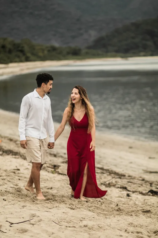 Casal caminhando de mãos dadas na areia, ela de vestido vermelho