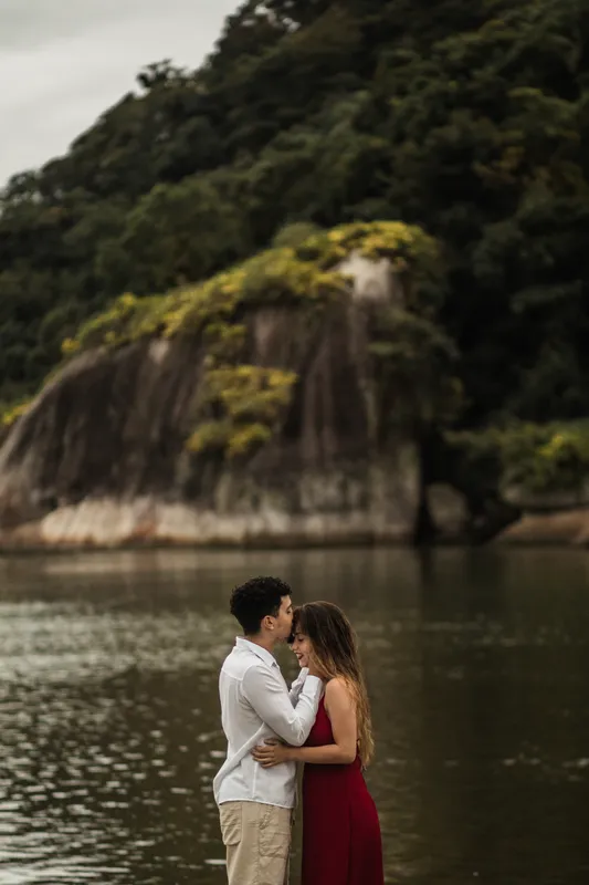 Casal abraçado na beira da água com uma pedra coberta de mata ao fundo