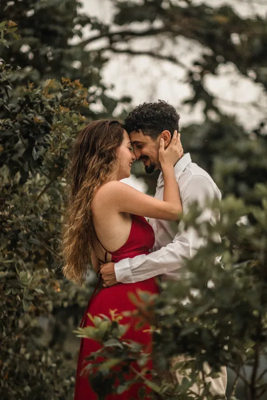 Casal de rosto colado entre folhagens, ela de vestido vermelho